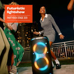 “A group of friends dancing outdoors next to a JBL PartyBox Stage 320 with its telescopic handle and wheels visible, displaying colorful LED lights.”