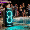 A JBL PartyBox Stage 320 portable speaker with glowing cyan LED accents stands on a stone patio beside a swimming pool at night, with a group of people gathered under a pergola in the background.