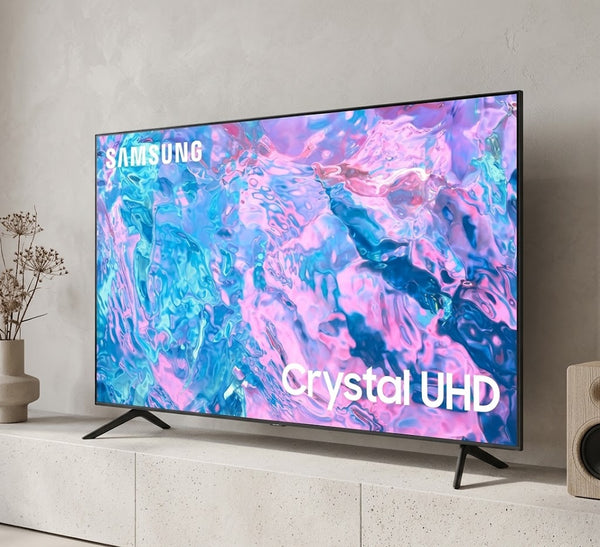 Samsung Crystal UHD television shown on a light stone console against a textured wall, with decorative dried flowers on the left and a speaker on the right.