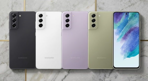 Five Samsung smartphones arranged side by side on a light marble surface, showing the back of the devices in black, white, lavender, sage green, and one with the screen on displaying a colorful wallpaper.